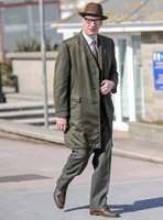 David Thewlis wearing glasses, tweed suit with overcoat and brown hat walking.