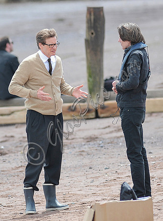 Colin Firth on Teignmouth beach, hands gesticulating, wearing beige cardigan talking to director James Marsh