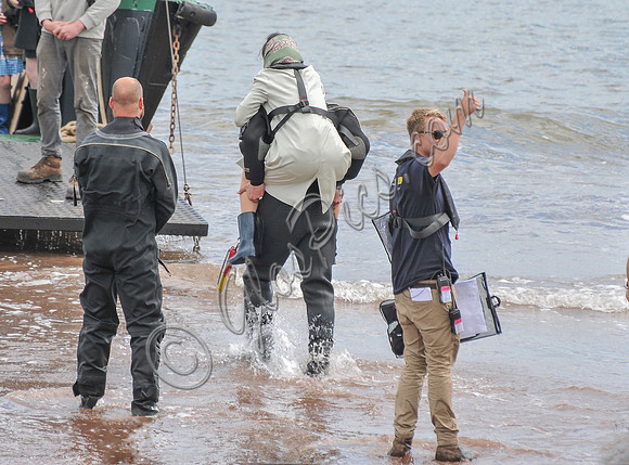 Rachel Weisz being piggy backed to  landing craft, wearing mackintosh, scarf and wellington boots