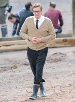 Colin Firth on Teignmouth beach, hands clenched loosely in front wearing beige cardigan, white shirt with brown tie, dark grey trousers and sailing boots