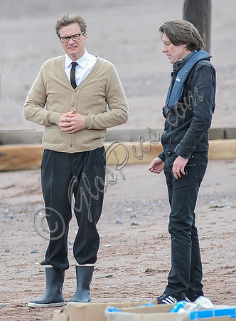 Colin Firth on Teignmouth beach, hands clenched loosely in front wearing beige cardigan talking to director James Marsh