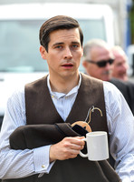 Rob James-Collier waring shirt and waistcoat looking directly at camera carrying jacket on clothes hanger and a white ceramic mug