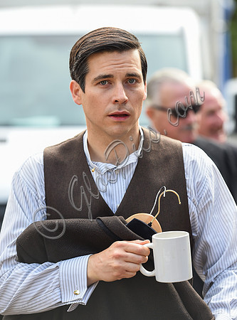 Rob James-Collier waring shirt and waistcoat looking directly at camera carrying jacket on clothes hanger and a white ceramic mug