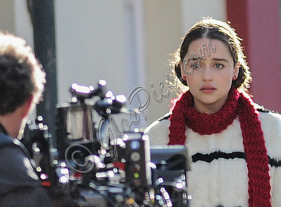Emilia Clarke on set wearing white with black stripe fur jacket, long red scarf, flowered skirt and yellow tights.