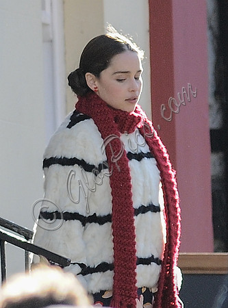 Emilia Clarke on set wearing white with black stripe fur jacket, long red scarf, flowered skirt and yellow tights.