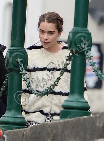 Emilia Clarke on set wearing white fur jacket with black stripes by green railings