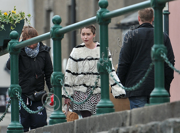 Emilia Clarke on set wearing white fur jacket with black stripes by green railings