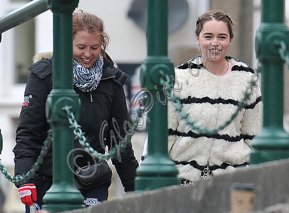 Emilia Clarke on set wearing white fur jacket with black stripes by green railings