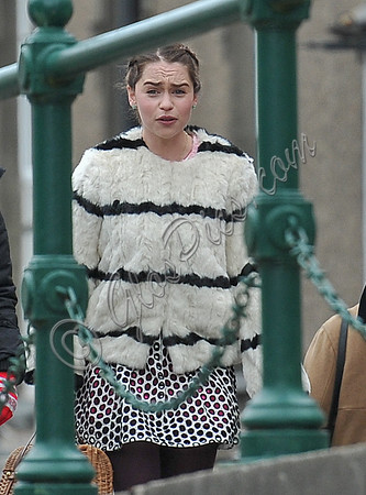 Emilia Clarke on set wearing white fur jacket with black stripes by green railings